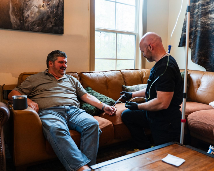 A man receives an IV infusion while seated on a brown leather couch in a living room.