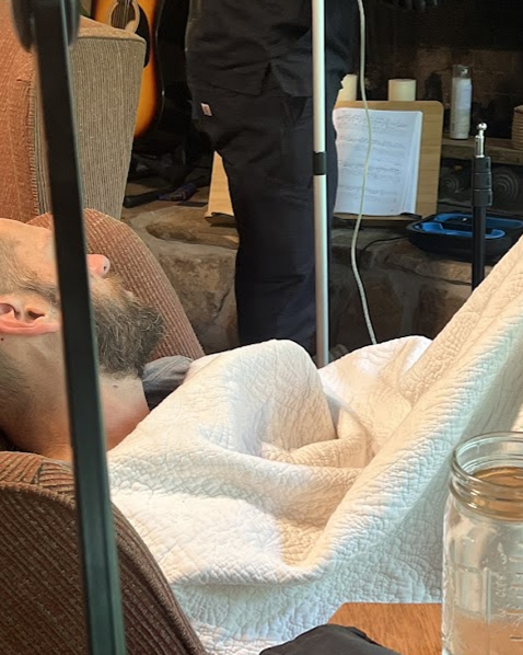 Man receiving IV fluids, lying down under a white blanket near a person holding up an IV pole indoors.