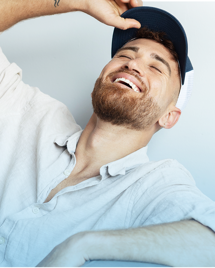 Man laughing, wearing a baseball cap and light-colored button-down shirt, head tilted back.