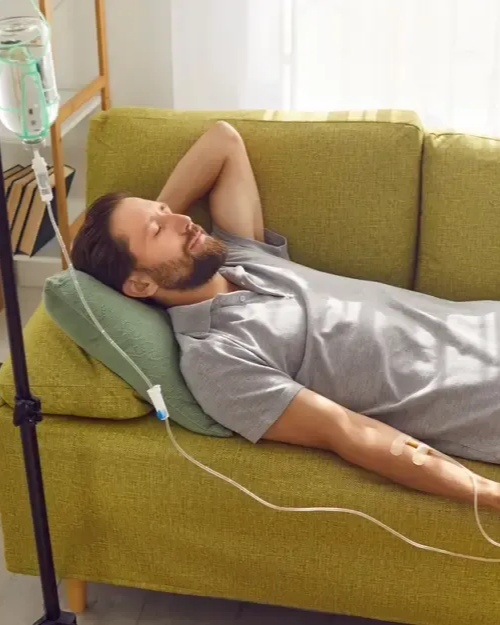 Man lying on a green couch, receiving an IV drip. He's relaxed, indoors, in a living room setting.