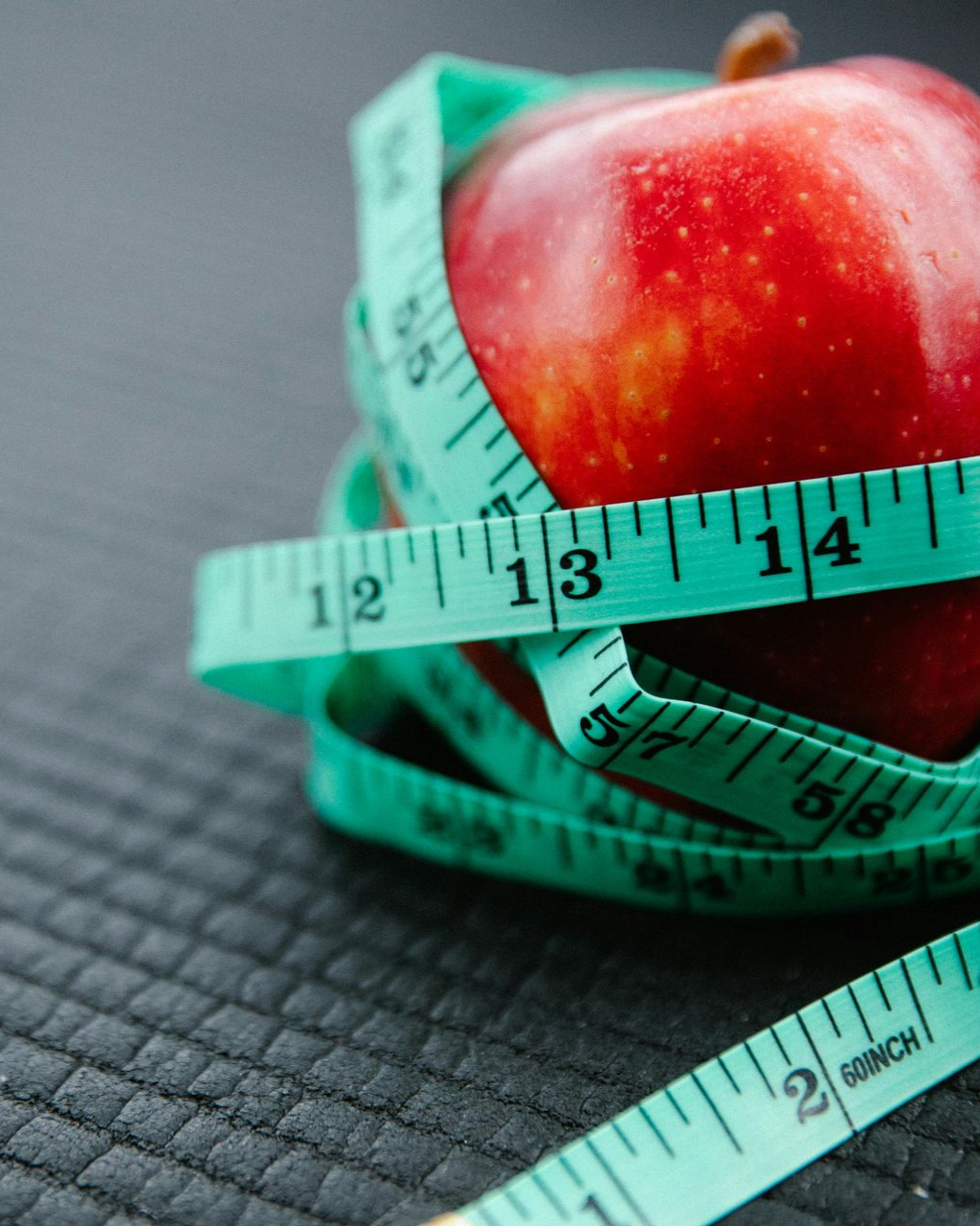 Red apple wrapped with a turquoise measuring tape on a textured, dark surface.