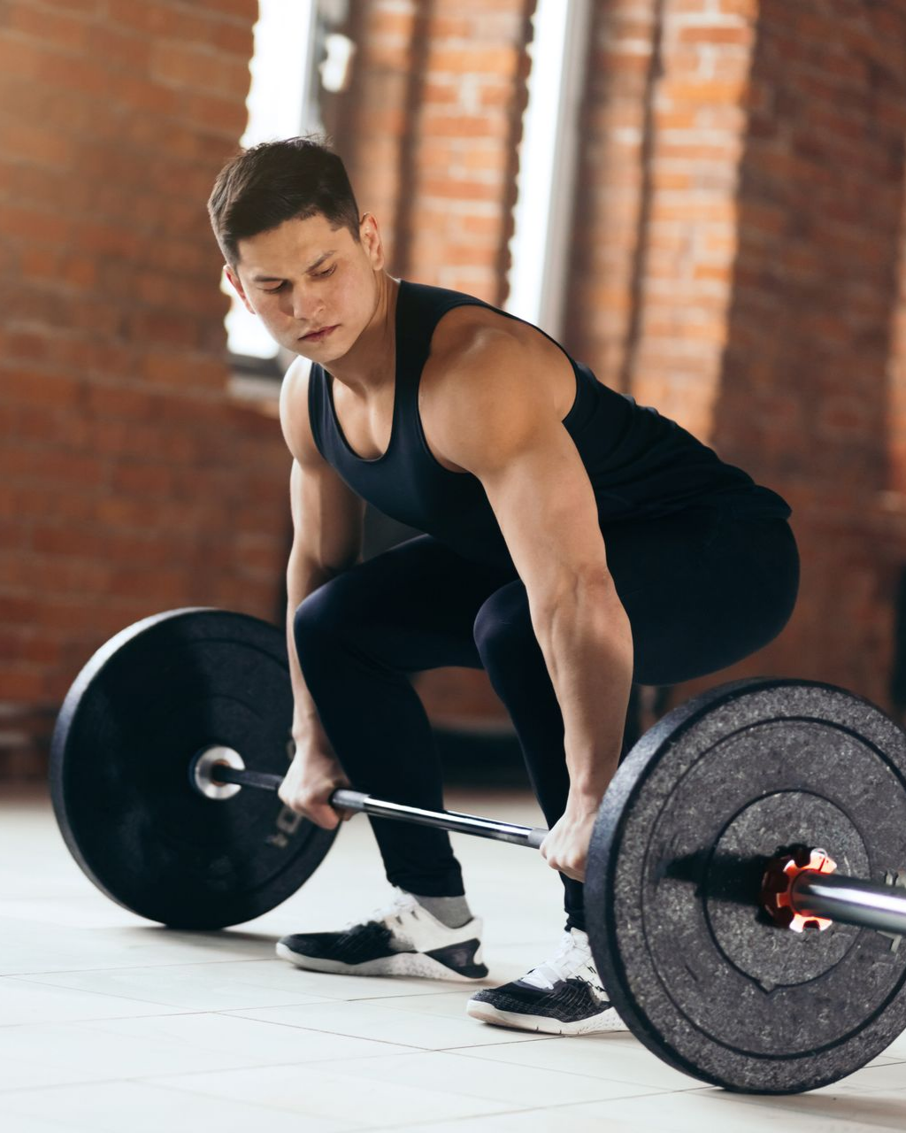 Man in gym lifting a barbell, preparing for a deadlift. He’s wearing black athletic clothing. Red brick wall.