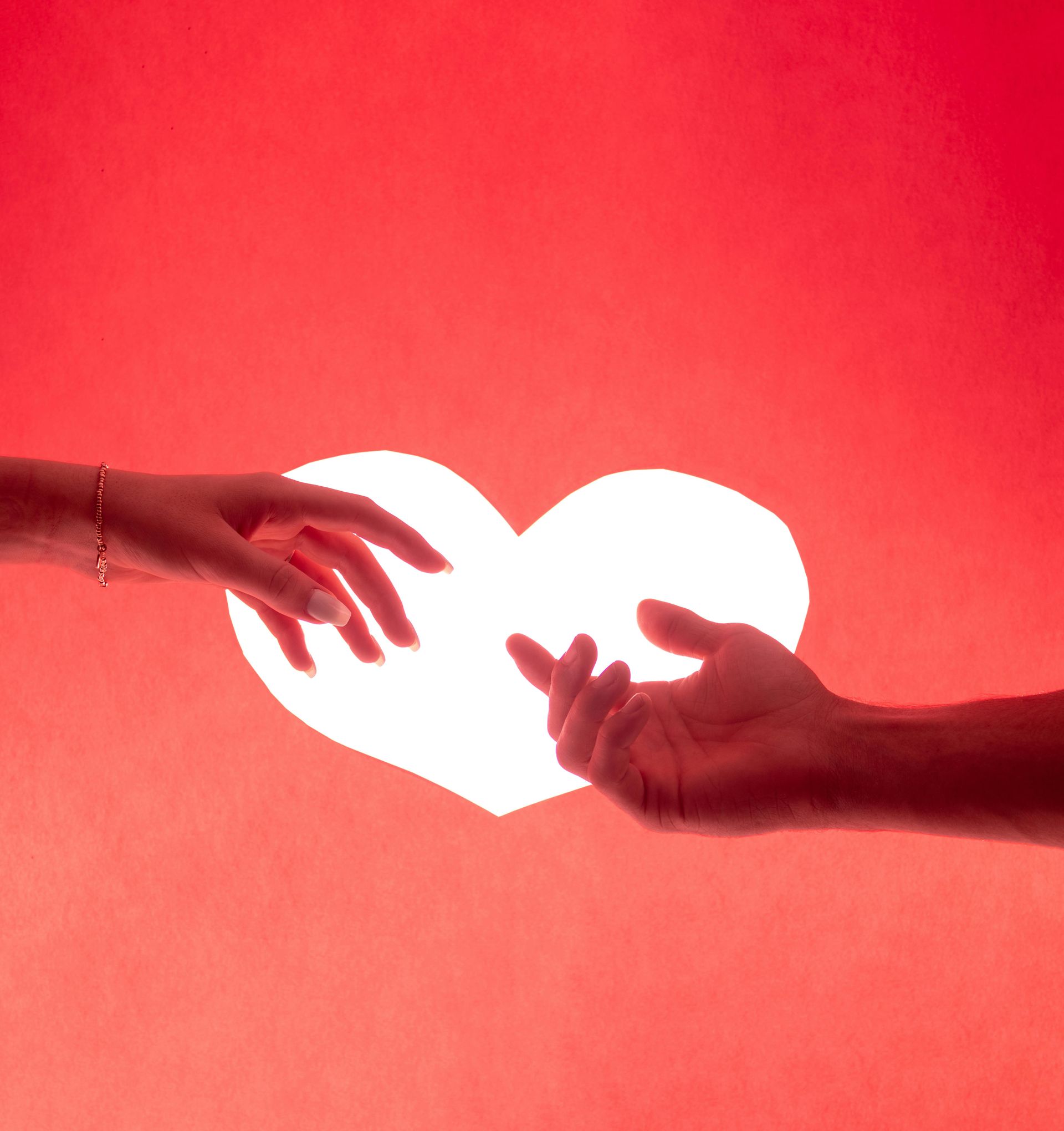 Due mani protese verso un cuore bianco luminoso su sfondo rosso.