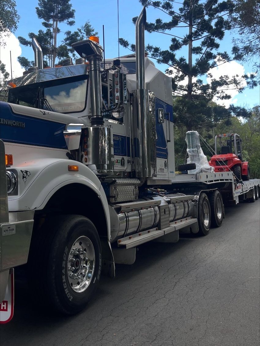 White and blue semi-truck with chrome details, pulling a flatbed trailer with a small red tractor, on a road — Supertilt Brisbane In Bracken Ridge, QLD