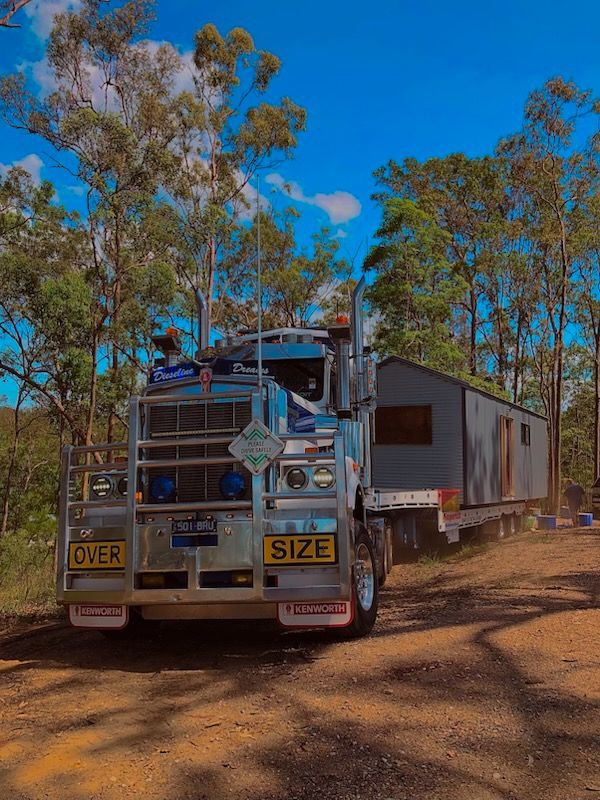 Semi-truck hauling a small building on a trailer down a dirt road, trees in the background, blue sky— Supertilt Brisbane In Bracken Ridge, QLD