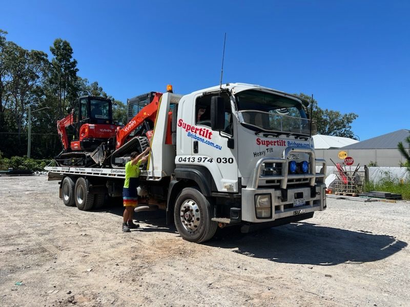 A white Supertilt flatbed truck carrying a small orange excavator on a gravel lot under a clear blue sky.