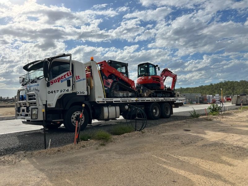 A white flatbed truck parked on a construction site, hauling a skid steer and a small excavator under a cloudy sky.