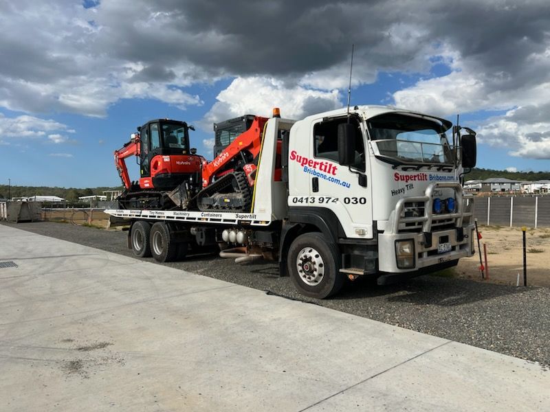 A white tow truck hauling an orange excavator and skid-steer loader on an outdoor lot under a cloudy sky.