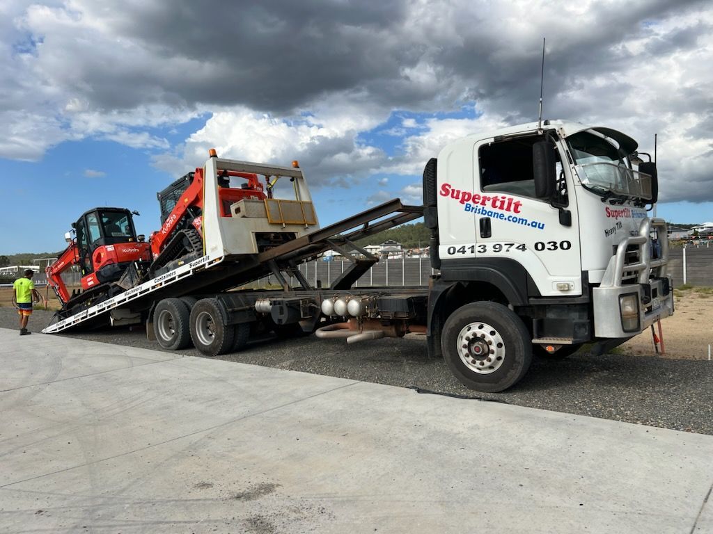 A white flatbed truck with an orange excavator on its tilted ramp, parked on a paved lot under a cloudy sky.