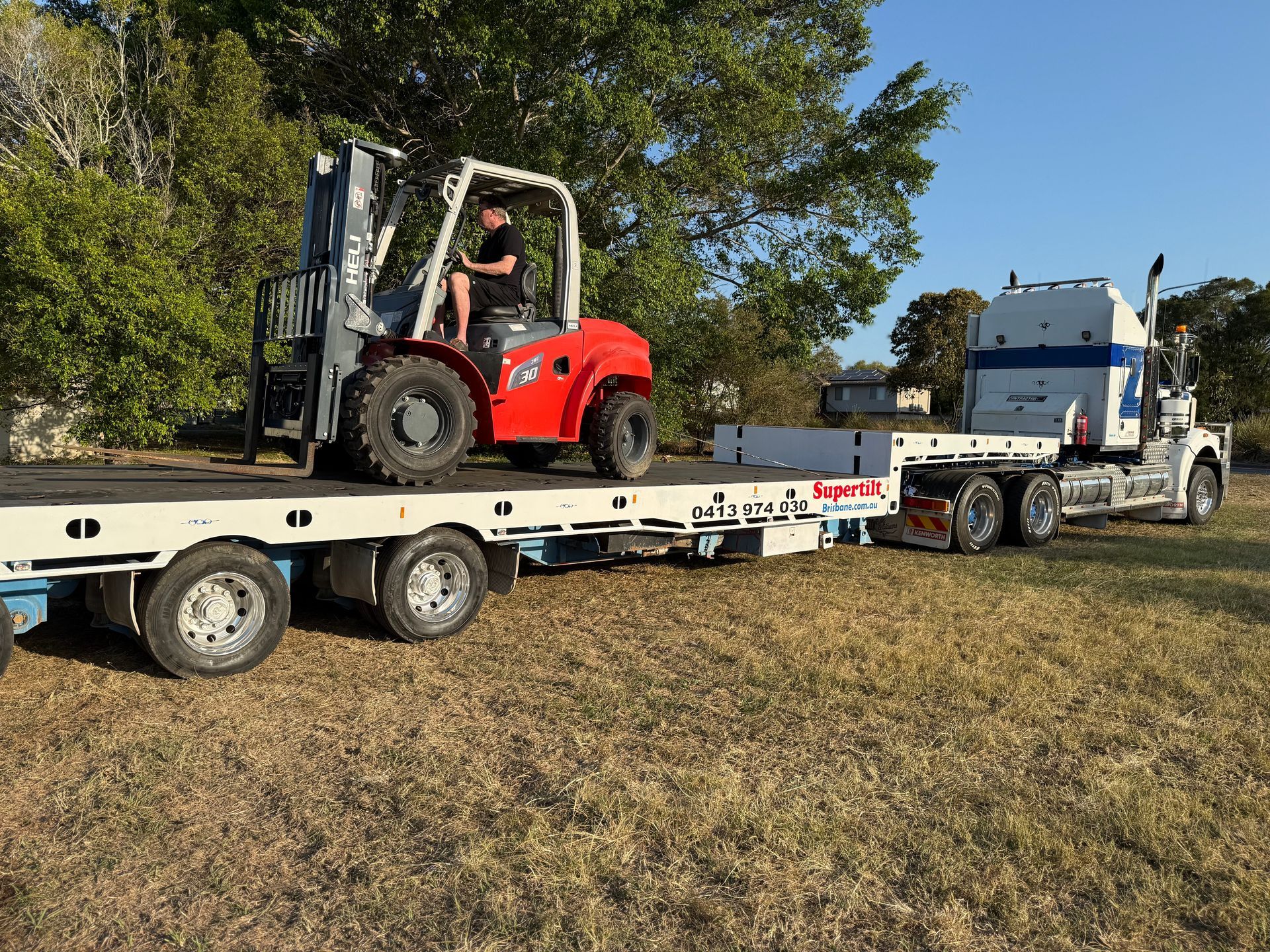 A red forklift on a flatbed trailer is being driven by a person. The truck is on grass — Supertilt Brisbane In Bracken Ridge, QLD