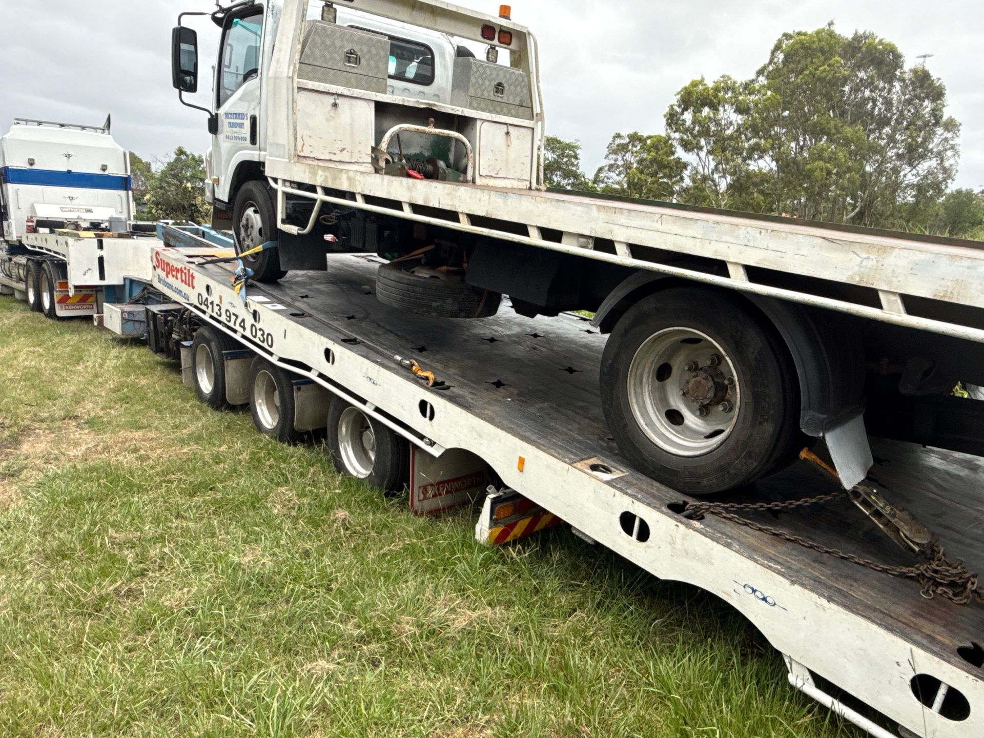 White truck on a low loader trailer parked on grass. Another truck is partially visible in the background. Cloudy sky — Supertilt Brisbane In North Lakes, QLD