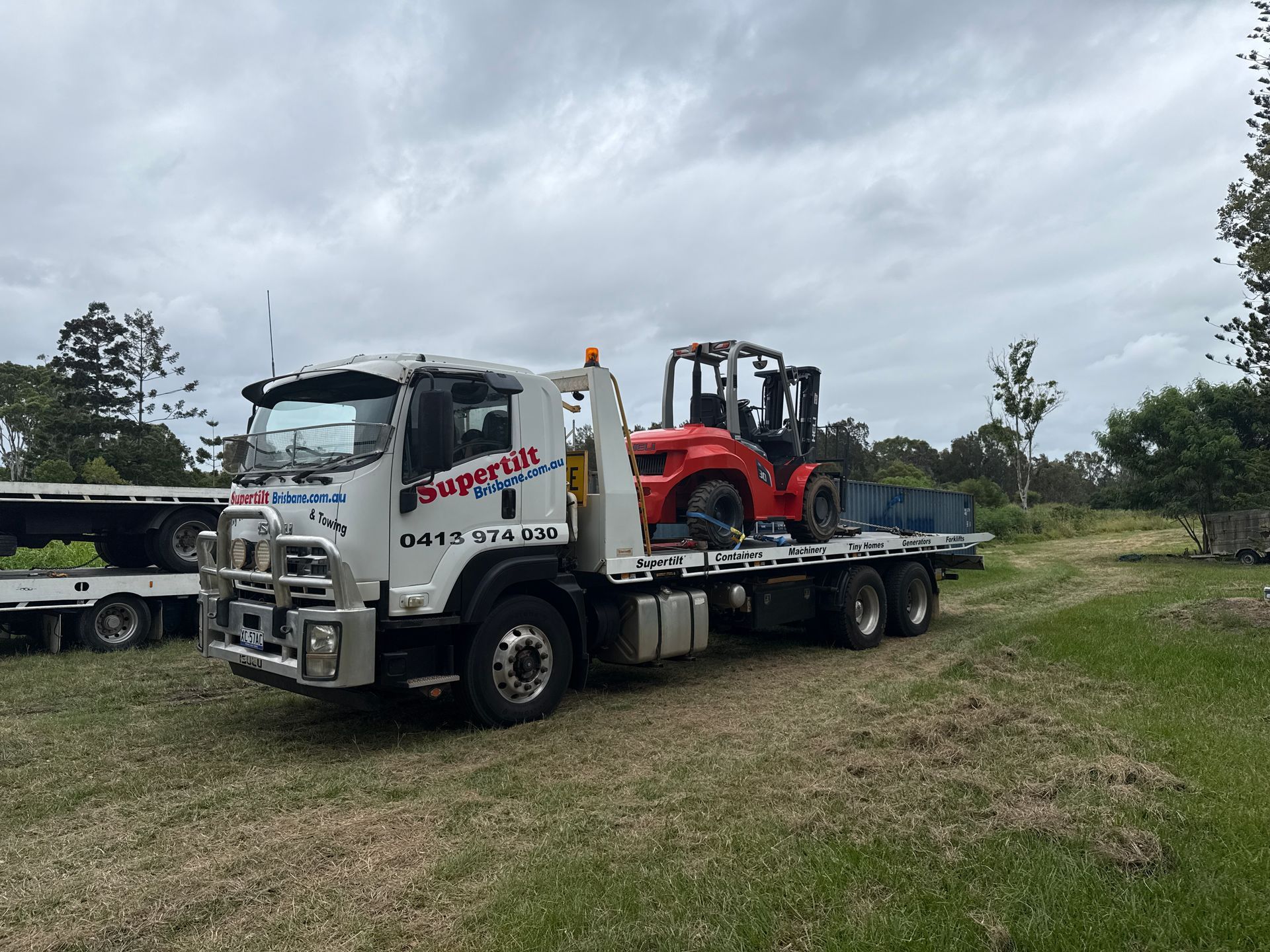 Tow truck carrying a red forklift on a flatbed in a grassy field under a cloudy sky — Supertilt Brisbane In North Lakes, QLD