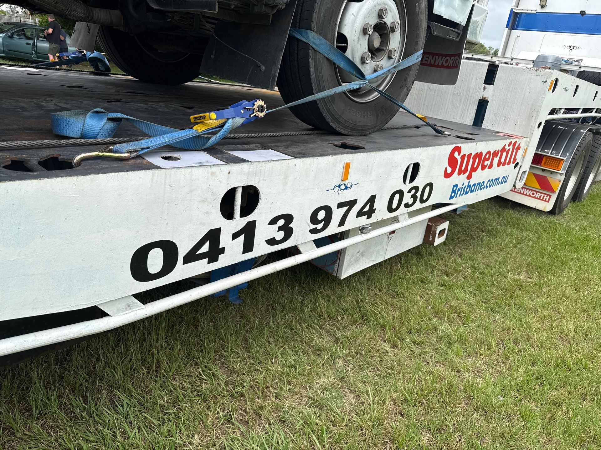 A flatbed tow truck with a vehicle secured, phone number on the side, on green grass — Supertilt Brisbane In North Lakes, QLD