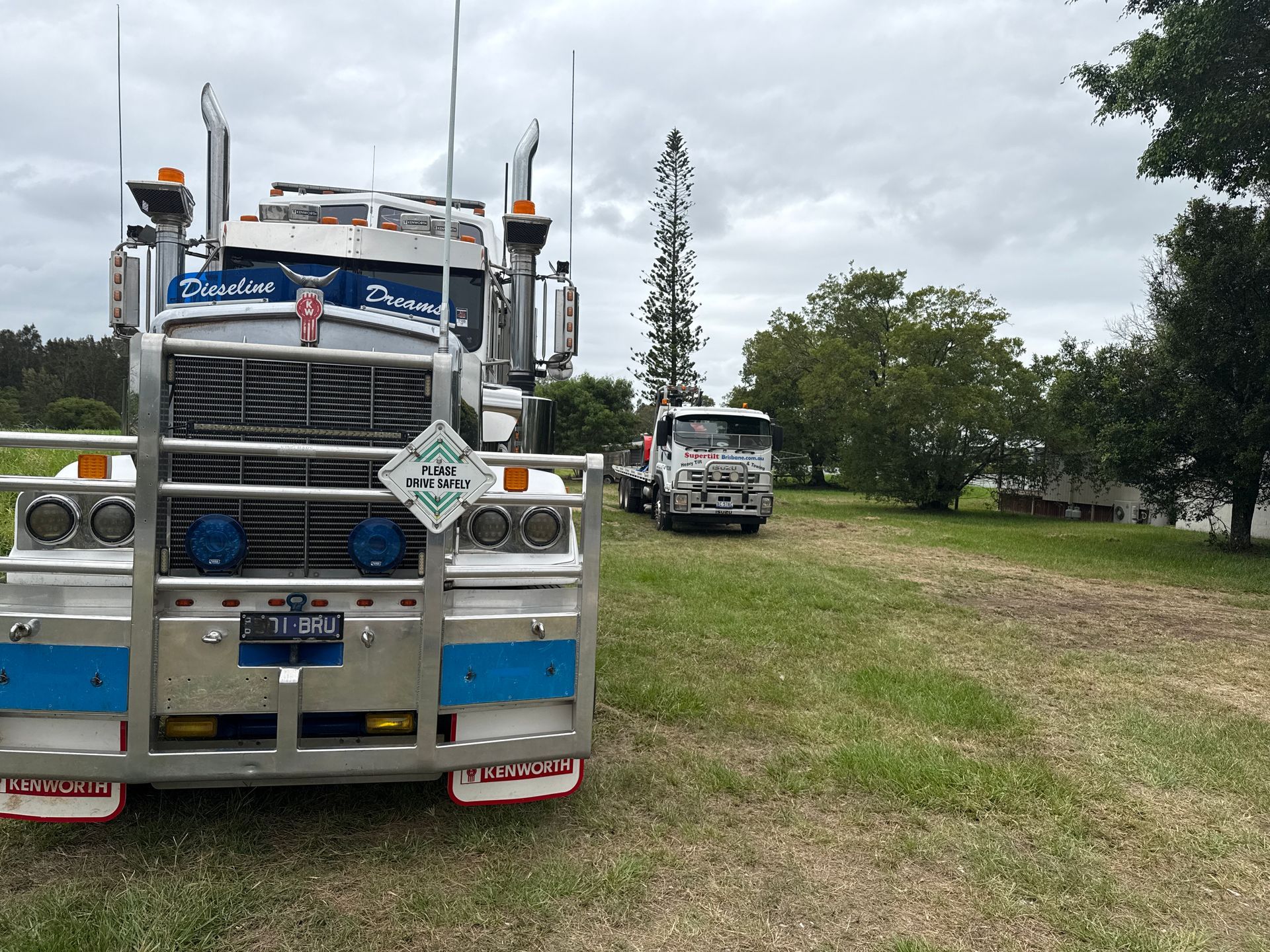 Two trucks on grass field; white Kenworth in foreground, other truck in background near trees and cloudy sky — Supertilt Brisbane In Bracken Ridge, QLD