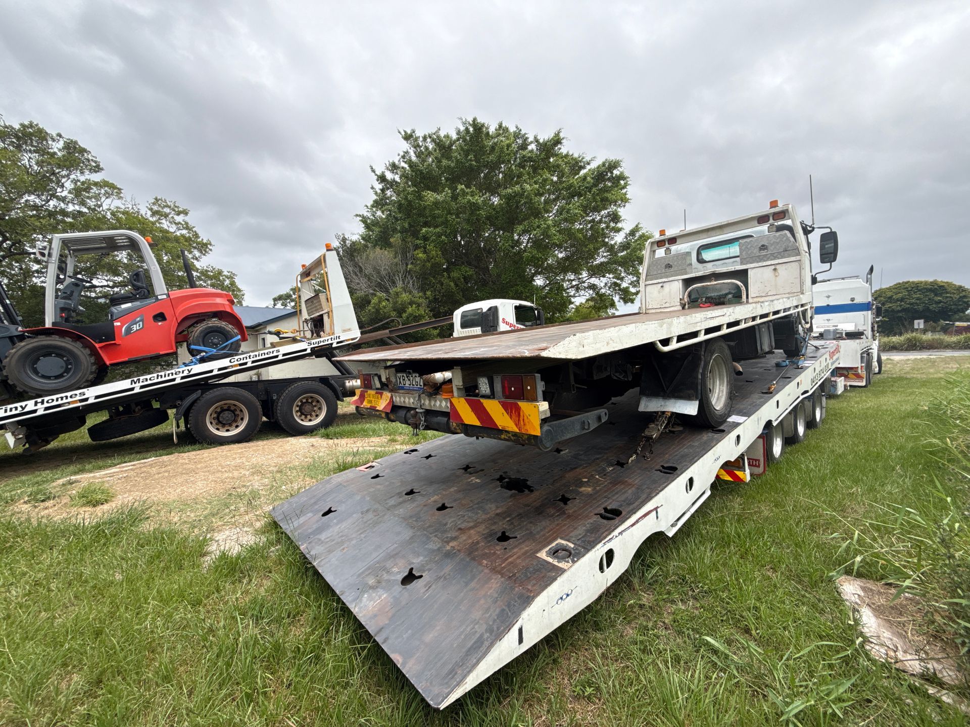 Two flatbed trucks carrying vehicles on grass. One truck has a small red forklift, the other has a white truck — Supertilt Brisbane In North Lakes, QLD