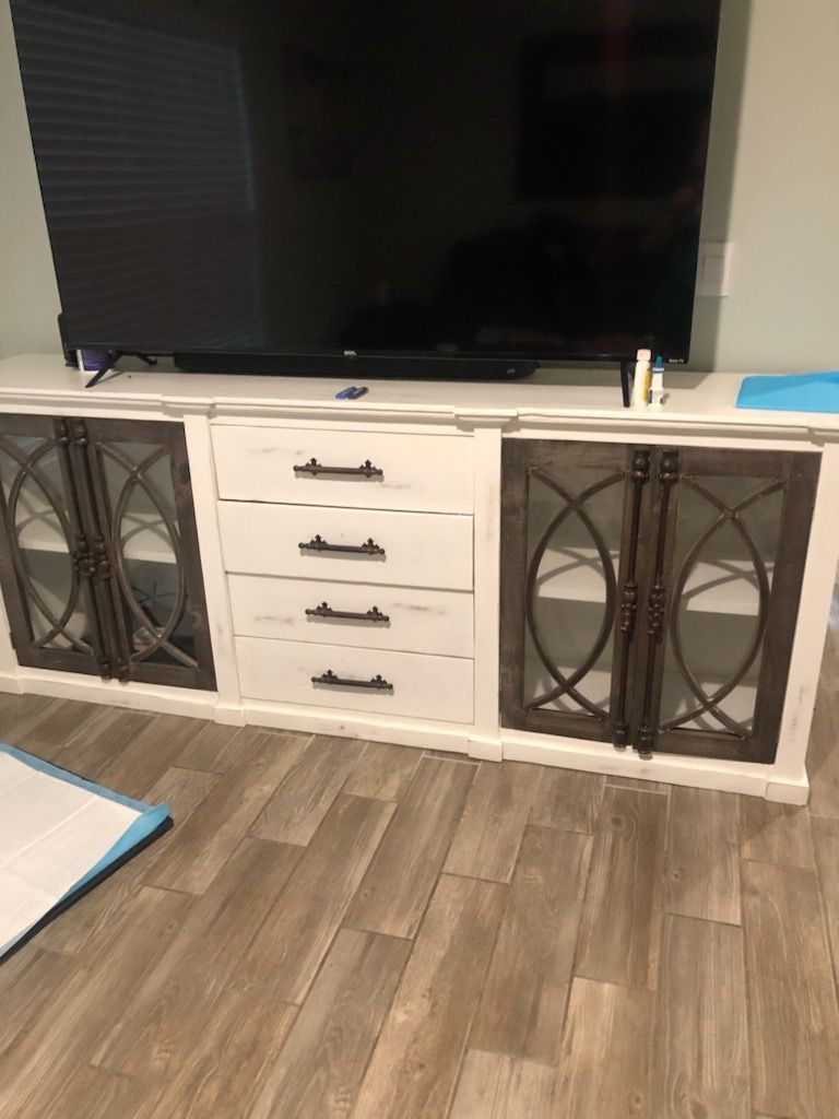 White TV console with drawers and glass-paneled doors, holding a large black TV, on a wood-look floor.