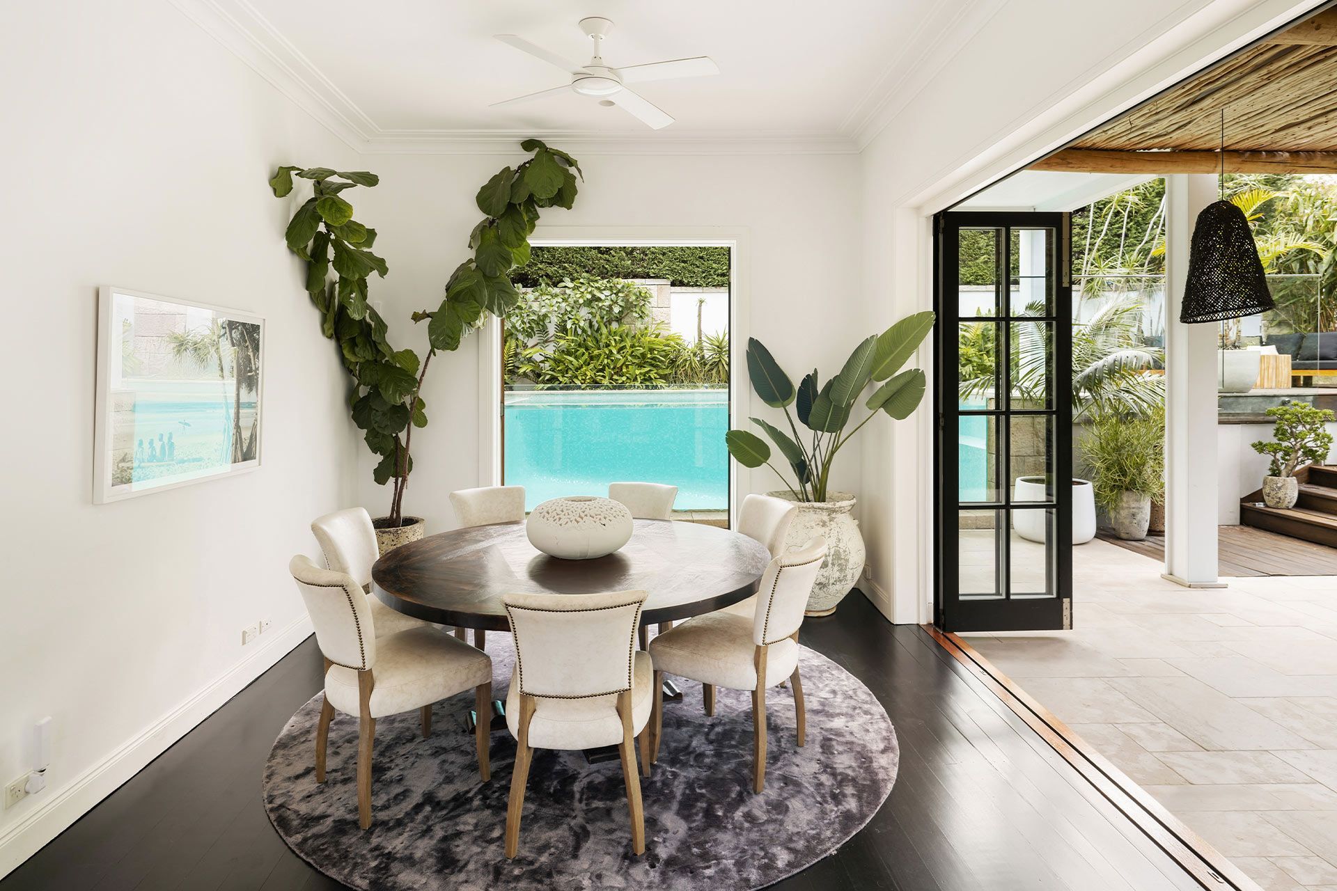 A dining room with a round table and chairs and a pool in the background. Cremorne