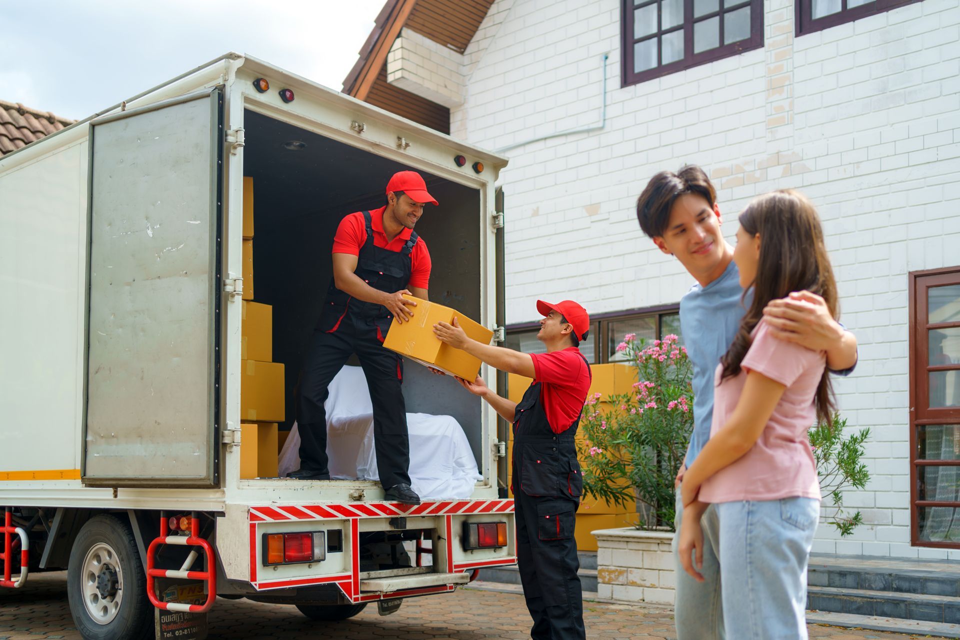Two movers in red uniforms unload boxes from a truck for a couple standing outside a house.