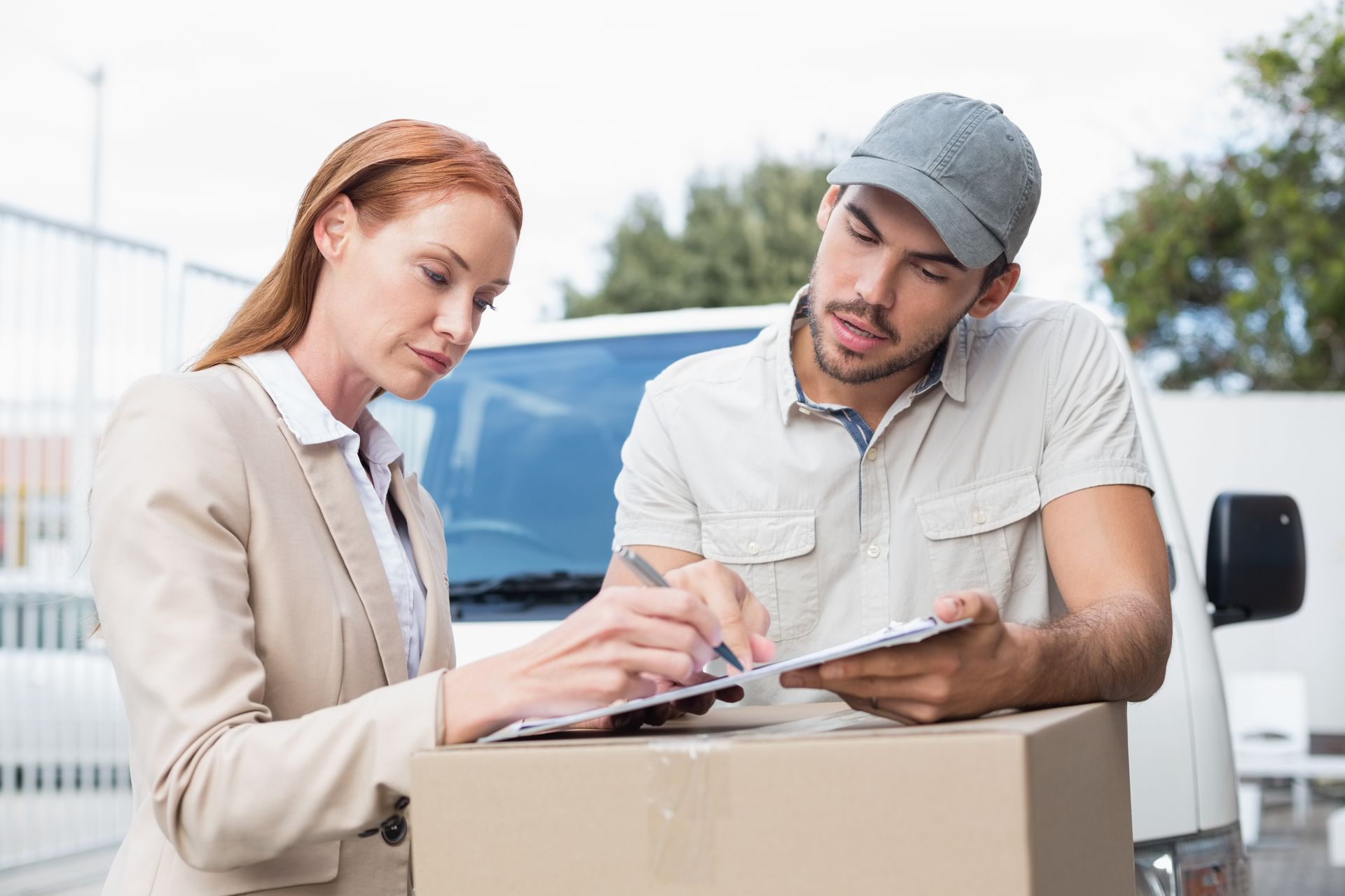 Woman signing a clipboard held by a delivery person next to a cardboard box and delivery van.
