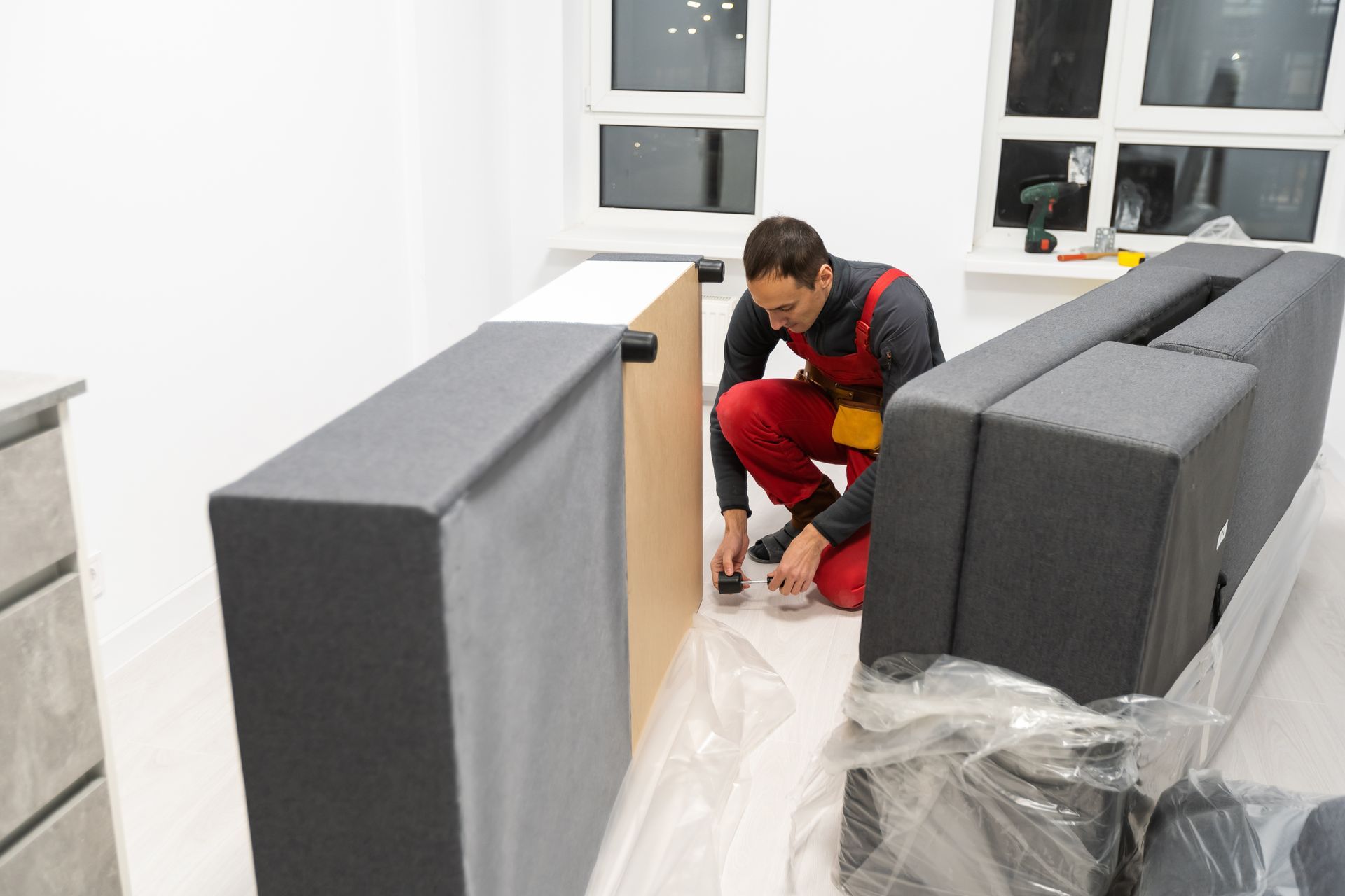 A person in red work overalls assembles furniture in a room with white walls and windows.