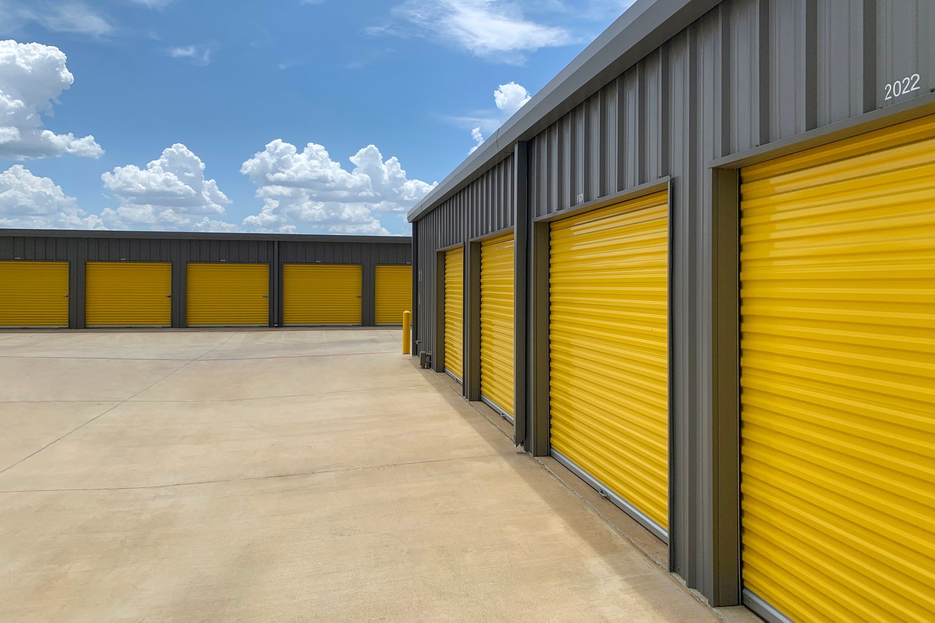 A row of storage units with bright yellow rolling doors under a blue sky with white clouds.