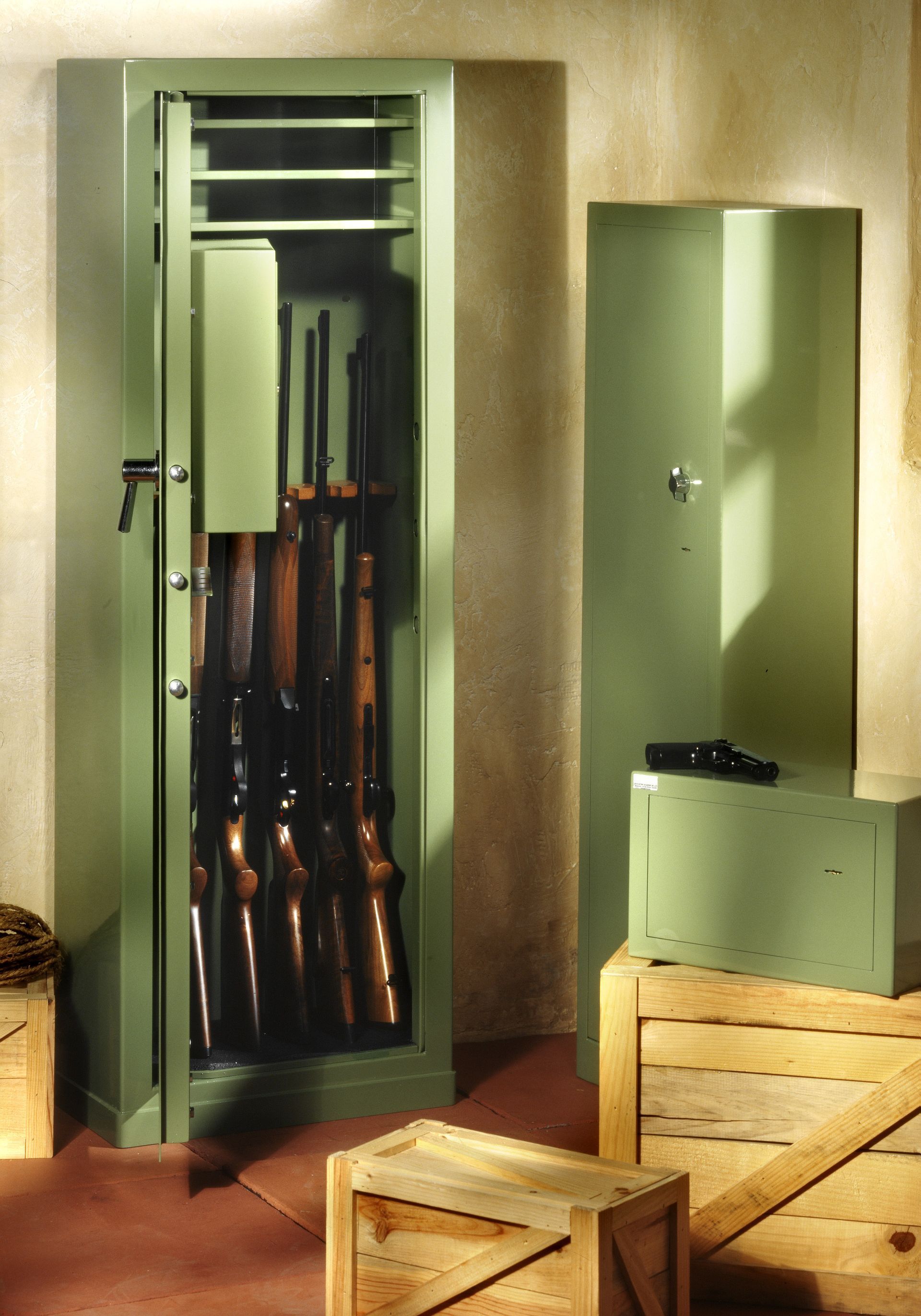 Green gun safe with rifles inside, standing next to a closed safe and a gun safe on a wooden box.