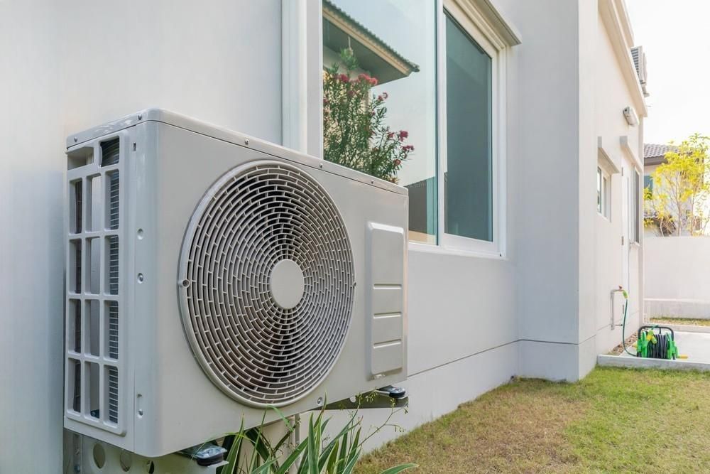 A white air conditioner is mounted on the side of a house next to a window.