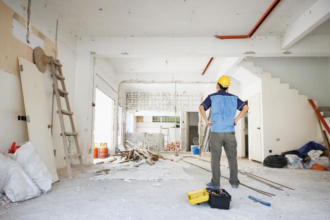 Construction worker in hard hat surveys a room under renovation; ladder, debris, and tools are visible.