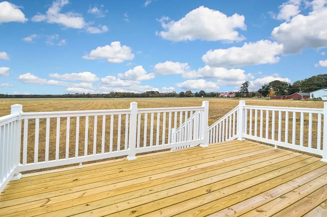 Wooden deck with white railing, overlooking a field under a blue sky with fluffy clouds.