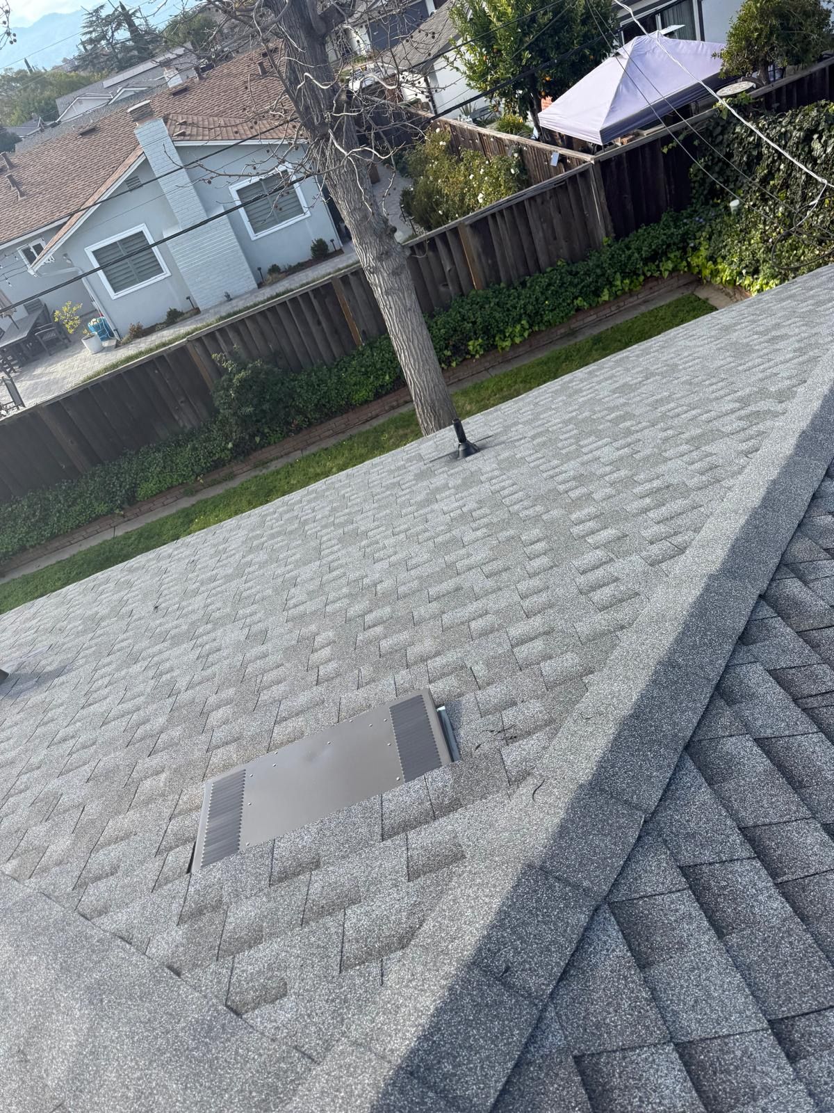 Slanted rooftop with gray shingles, a small vent, and a backyard fence and tree in the background