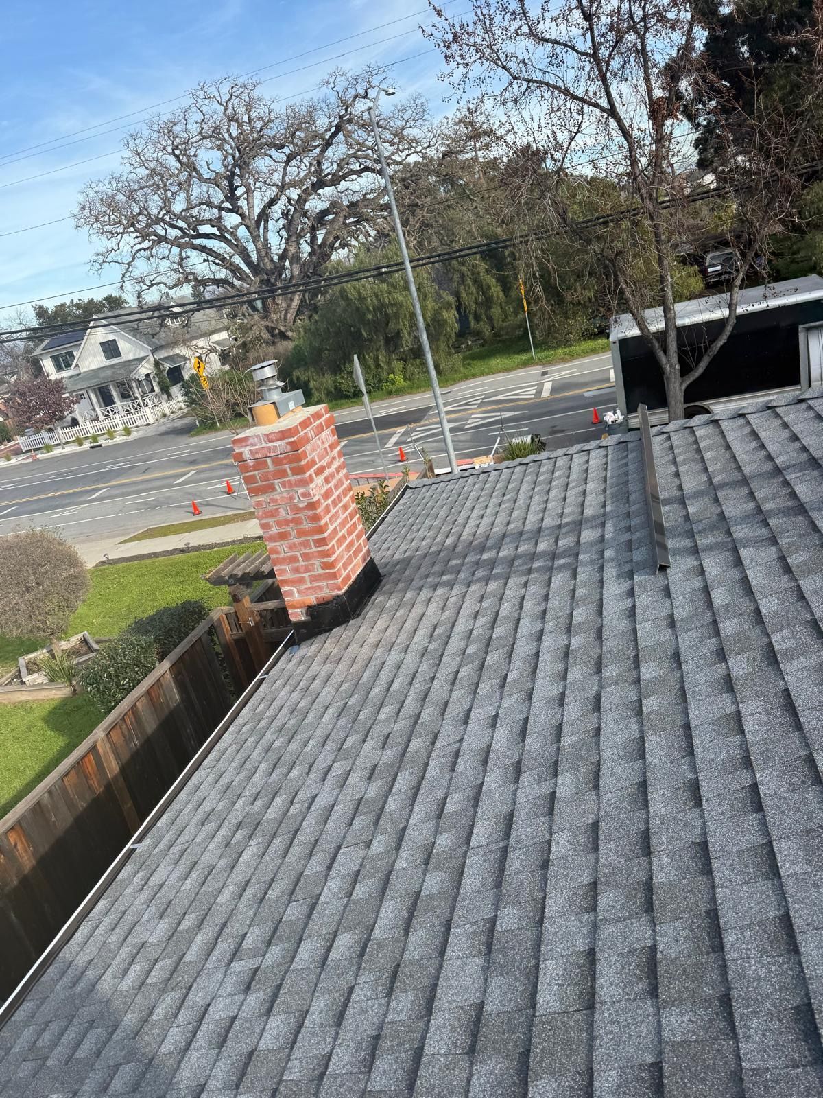 Rooftop view with gray shingles, a brick chimney, trees, and a road in the background