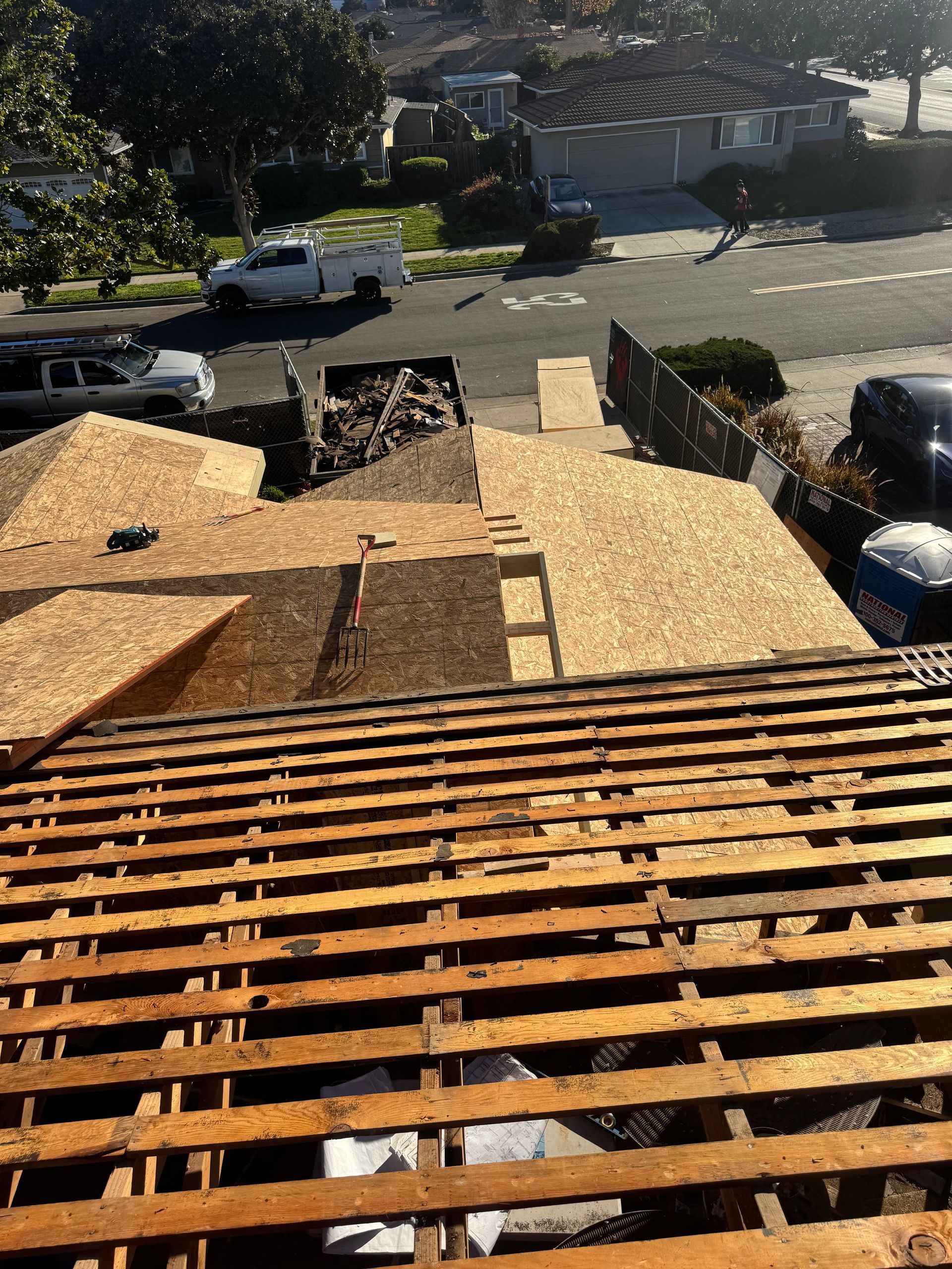 Roofer’s view of a residential street with exposed wooden roof framing and plywood sheathing in the foreground