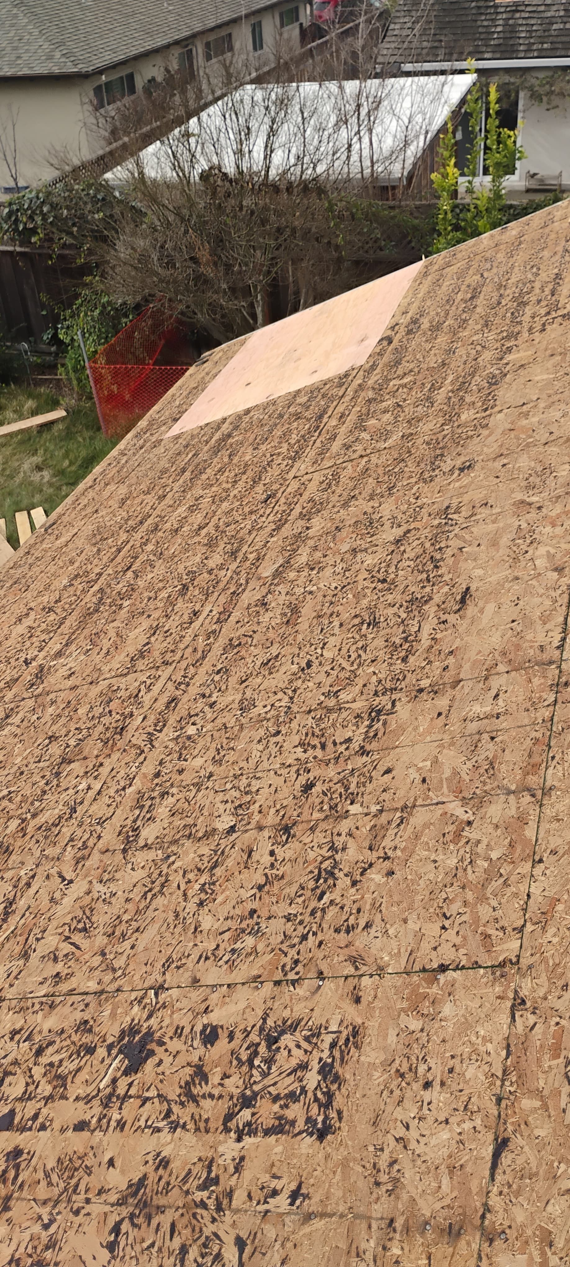 Close-up of a sloped roof covered in tan roofing material, with nearby houses and trees in the background.