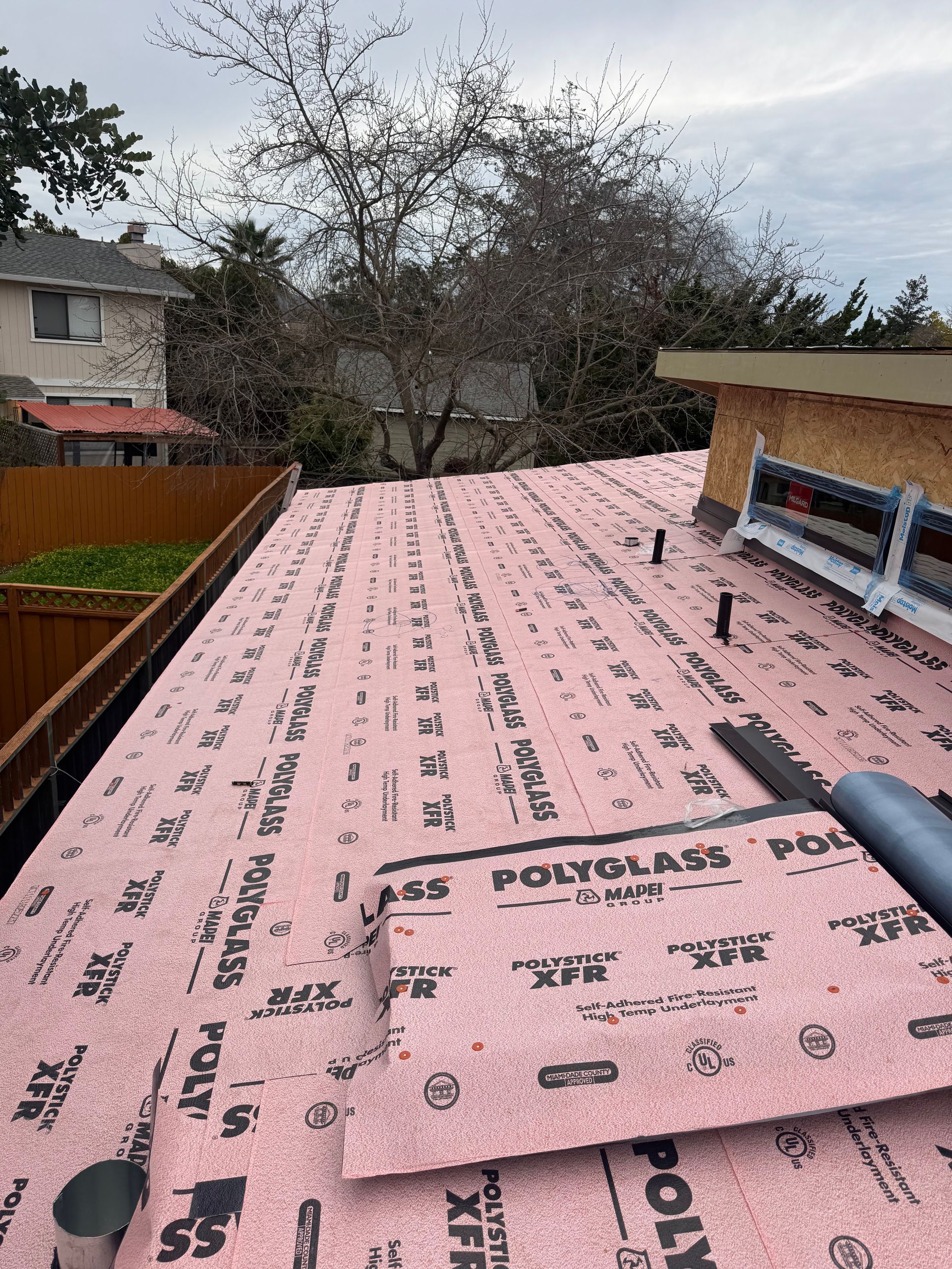 Roof under construction with pink underlayment, chimney vent, and trees in the background