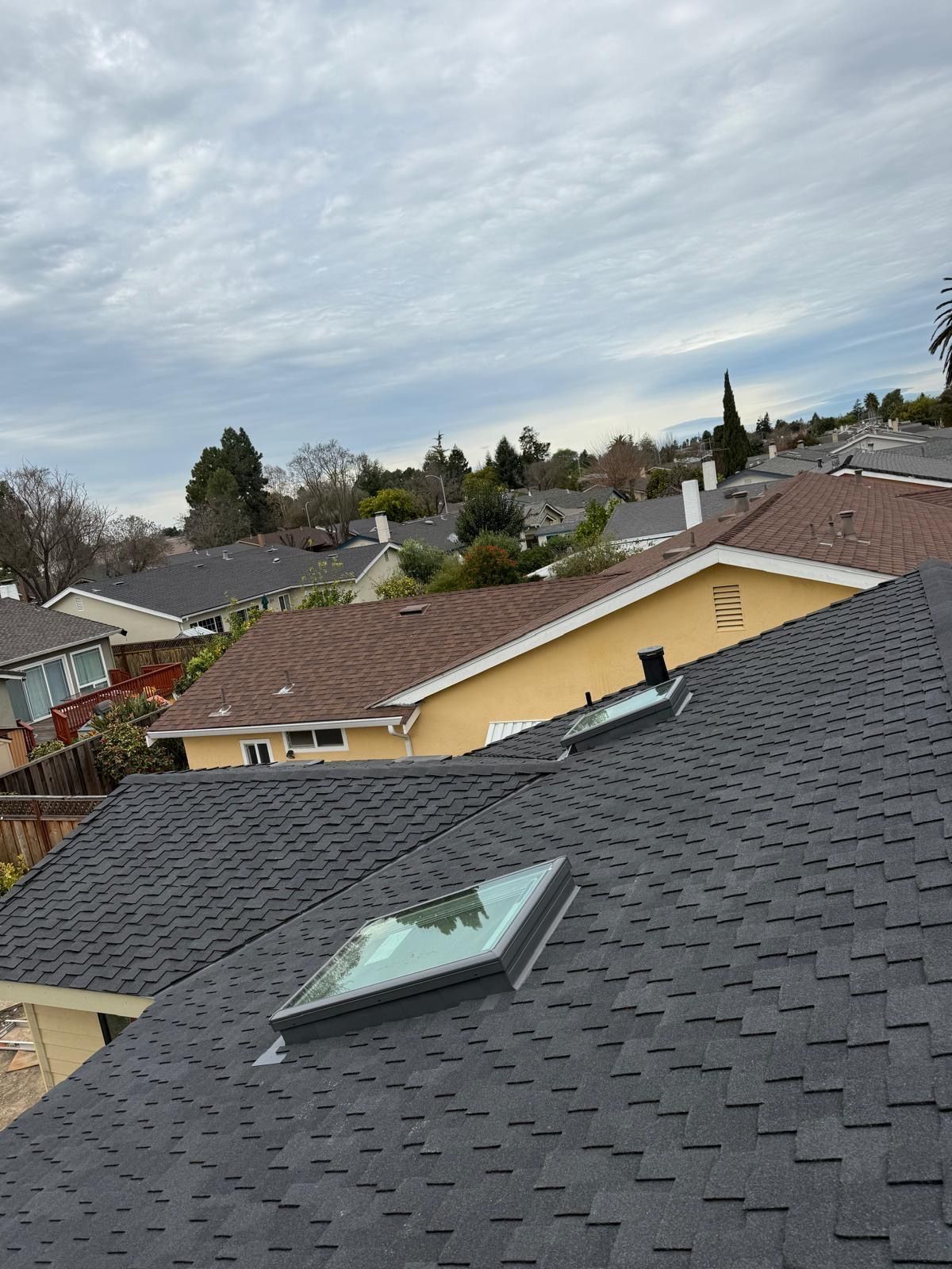 Suburban rooftops under a cloudy sky, with a skylight on the nearest dark shingle roof.
