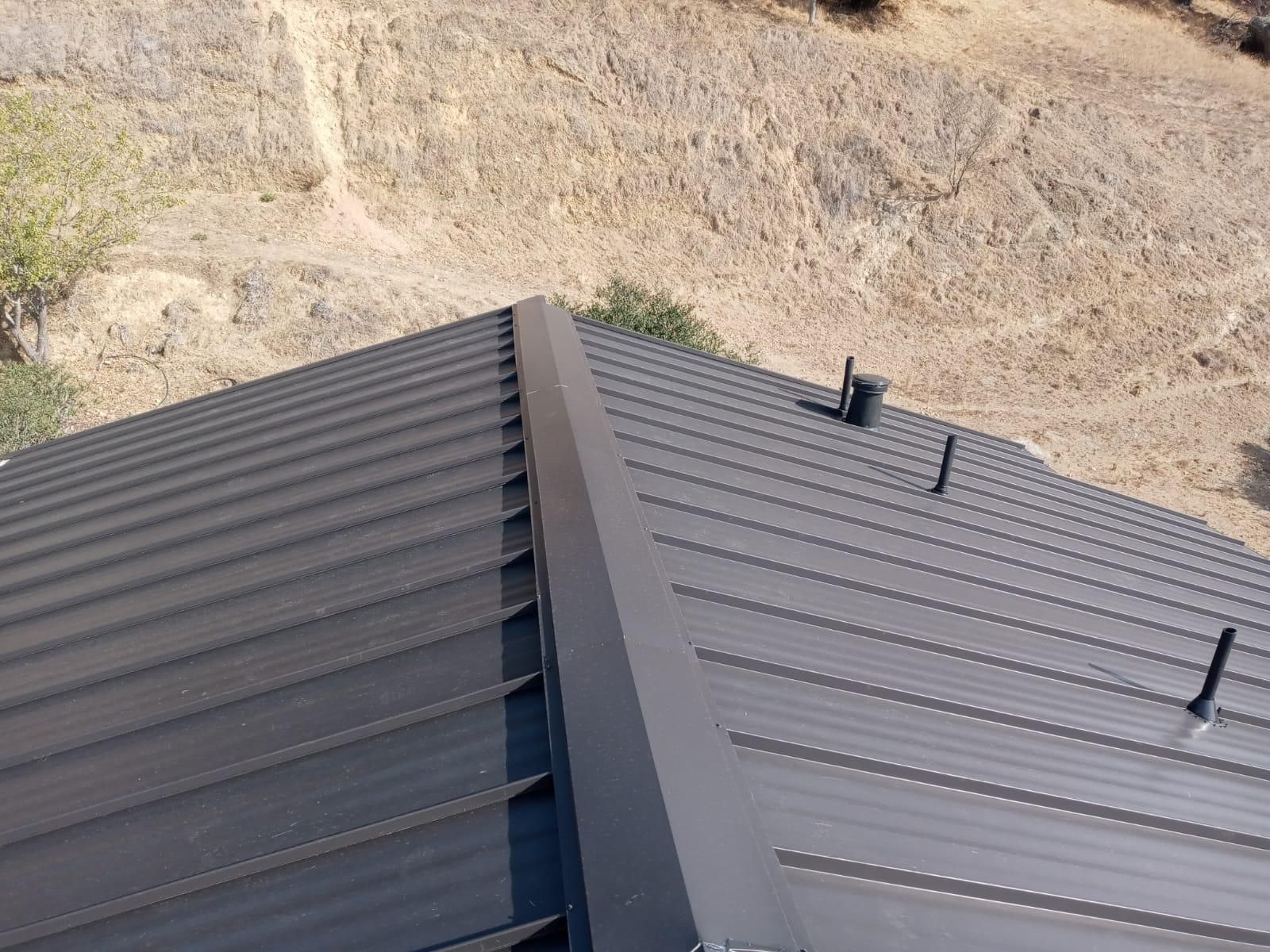 Metal roof on a house beside a dry, rocky hillside, with vents and a central ridge.