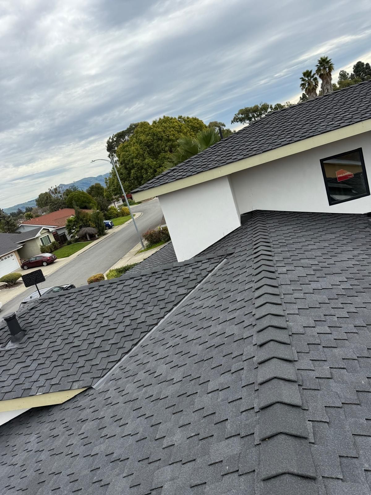 Rooftop with dark gray shingles overlooking a suburban neighborhood under a cloudy sky.