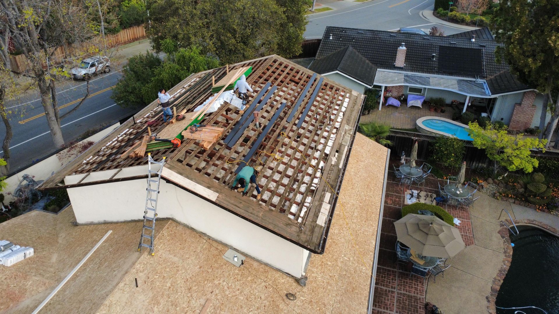 Aerial view of a roof under repair with exposed wooden battens and a nearby house, trees, and pool.