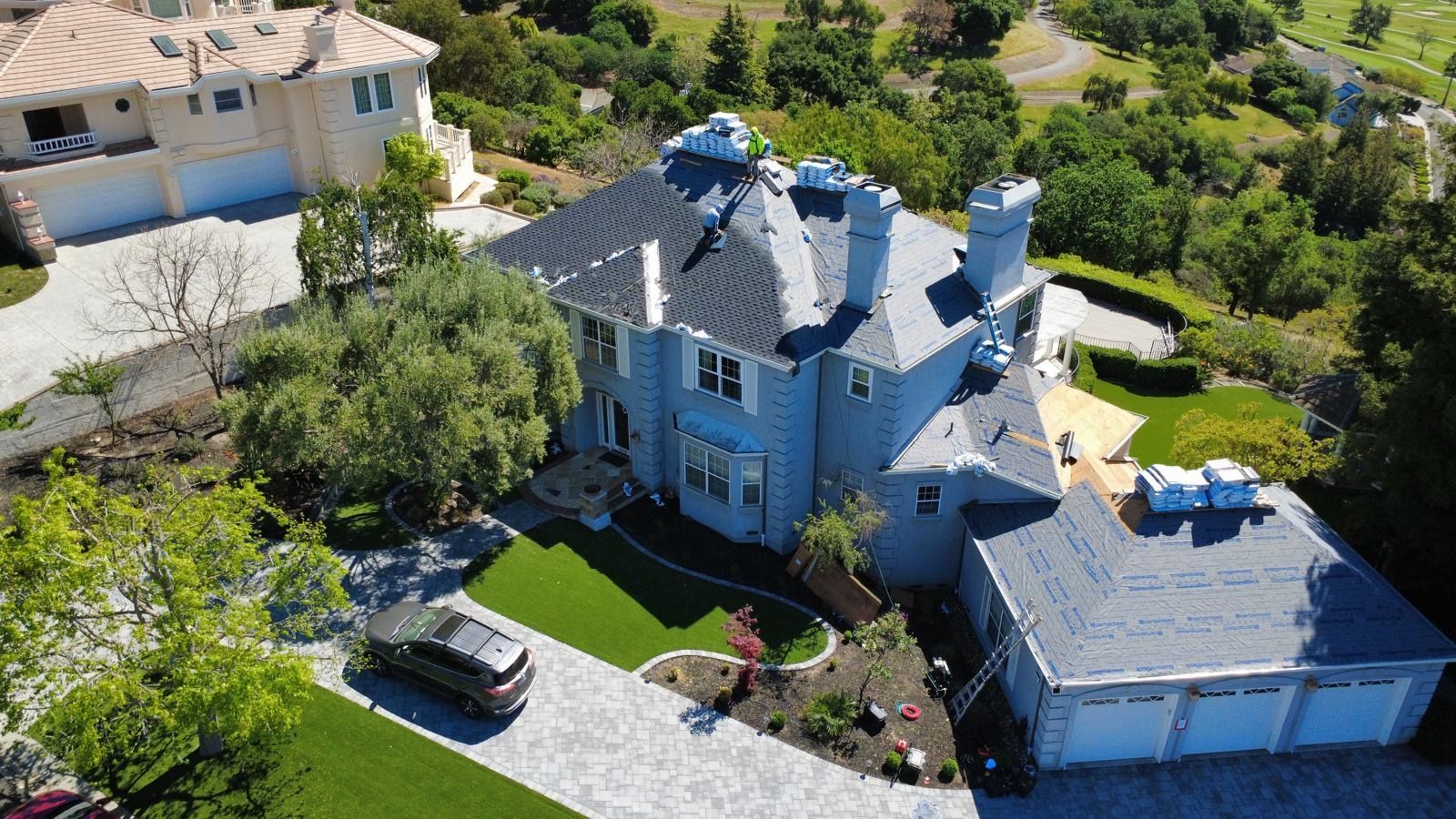Aerial view of a blue house with a driveway, garage, and lush green trees in a suburban neighborhood