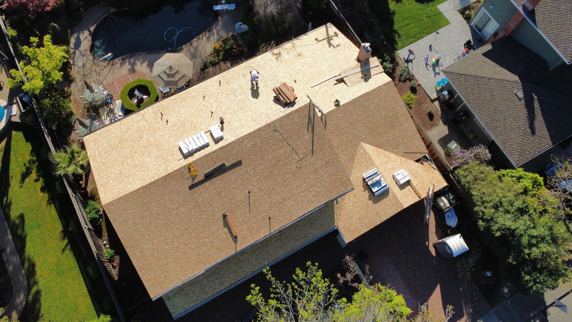 Aerial view of a beige-roofed house with rooftop vents, surrounded by trees and neighboring homes.