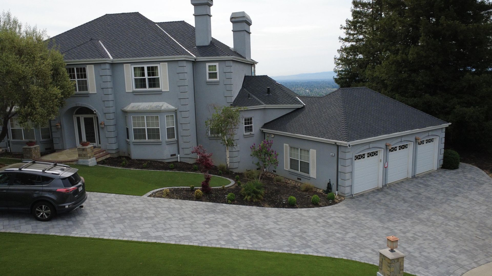 Large gray stone house with a garage, cobblestone driveway, and parked black SUV on a hillside.