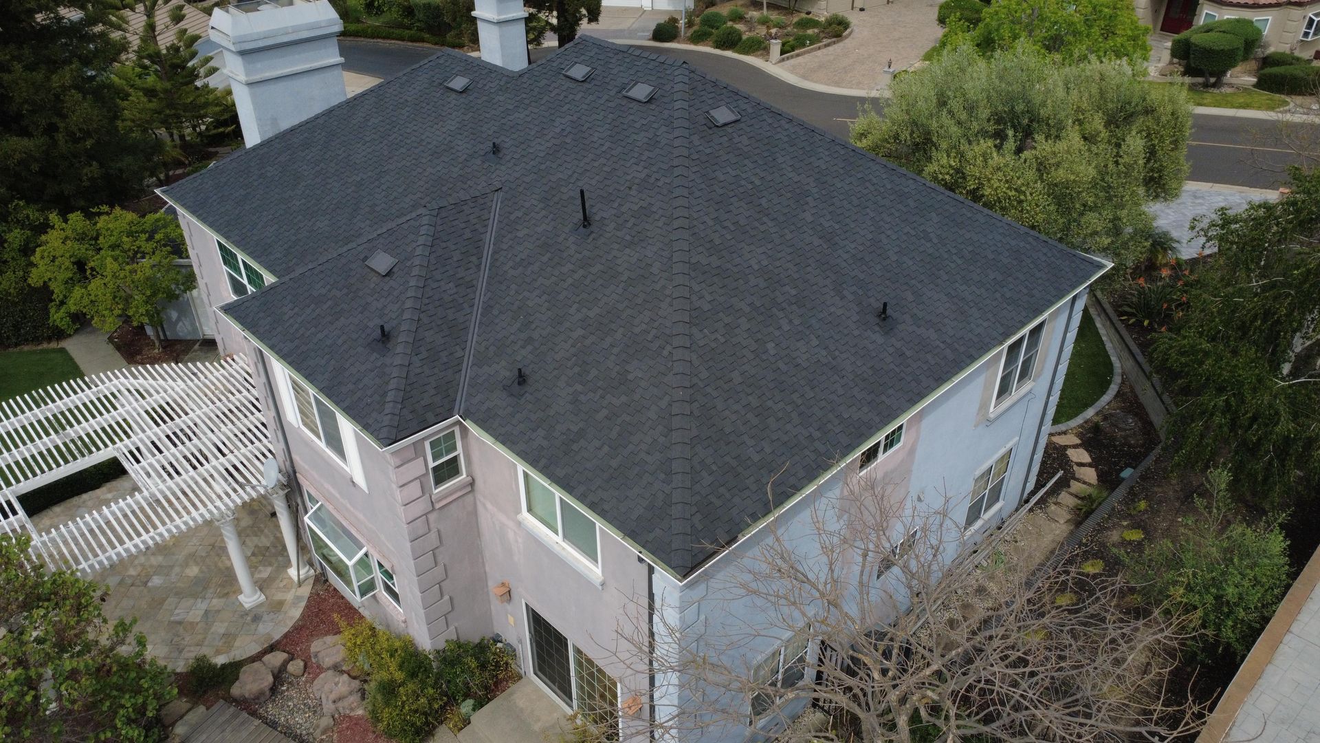 Aerial view of a gray-roofed house with light siding, surrounded by trees and neighboring homes.