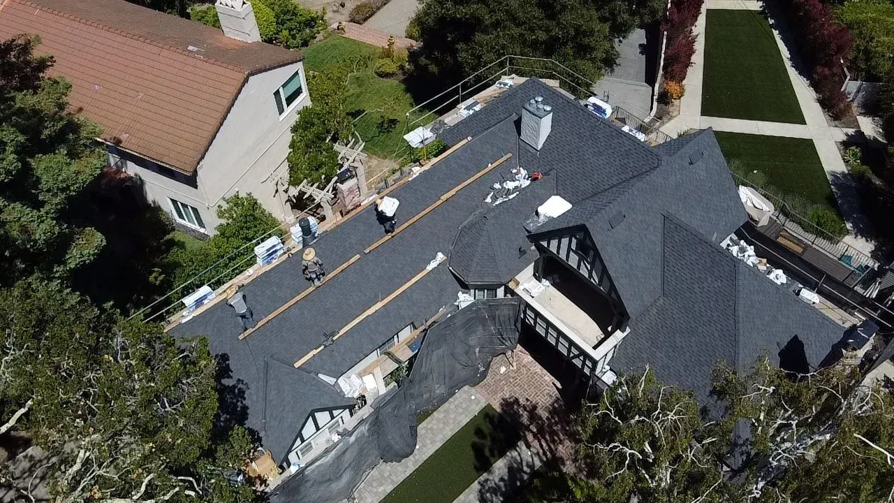 Aerial view of a house with a dark gray roof under repair, with workers and materials on top.