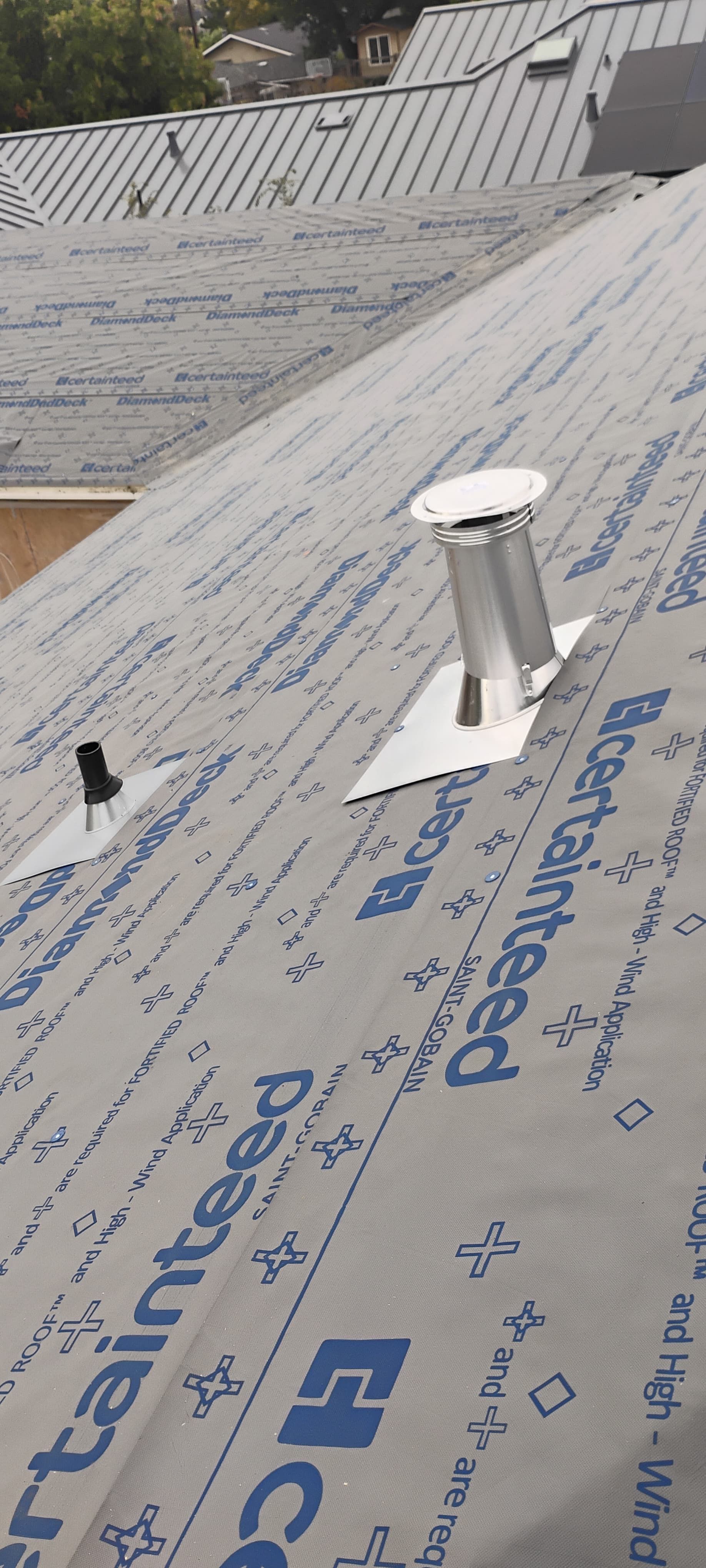 Roof under construction covered with blue underlayment and a metal vent pipe under a cloudy sky