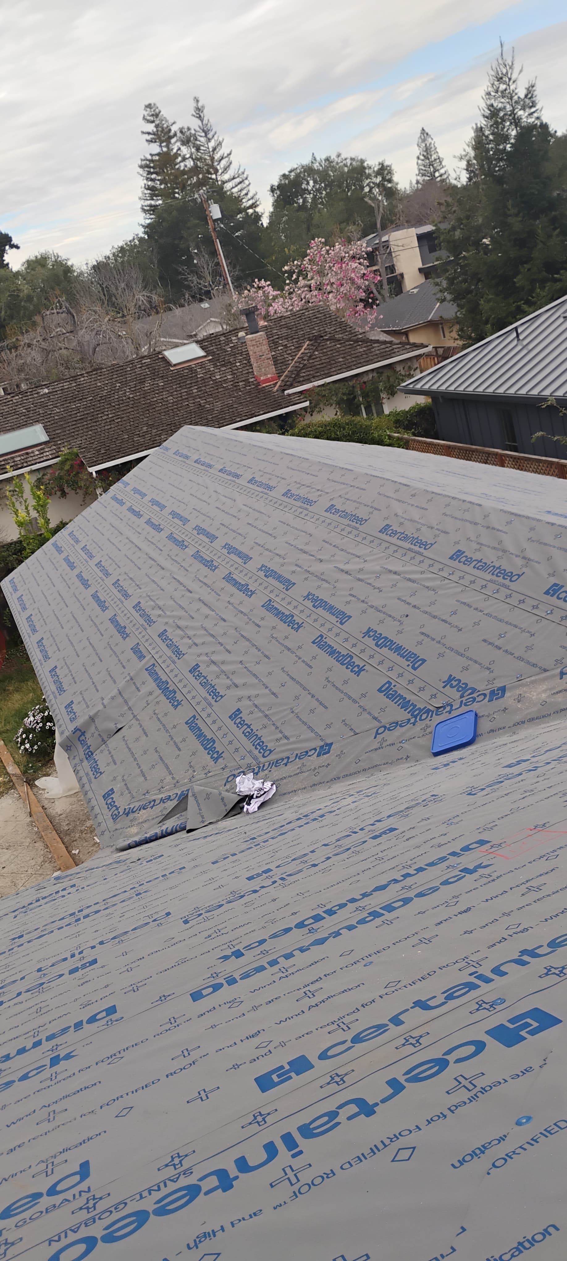 Roof under construction with gray underlayment, surrounded by trees and neighboring houses under a cloudy sky