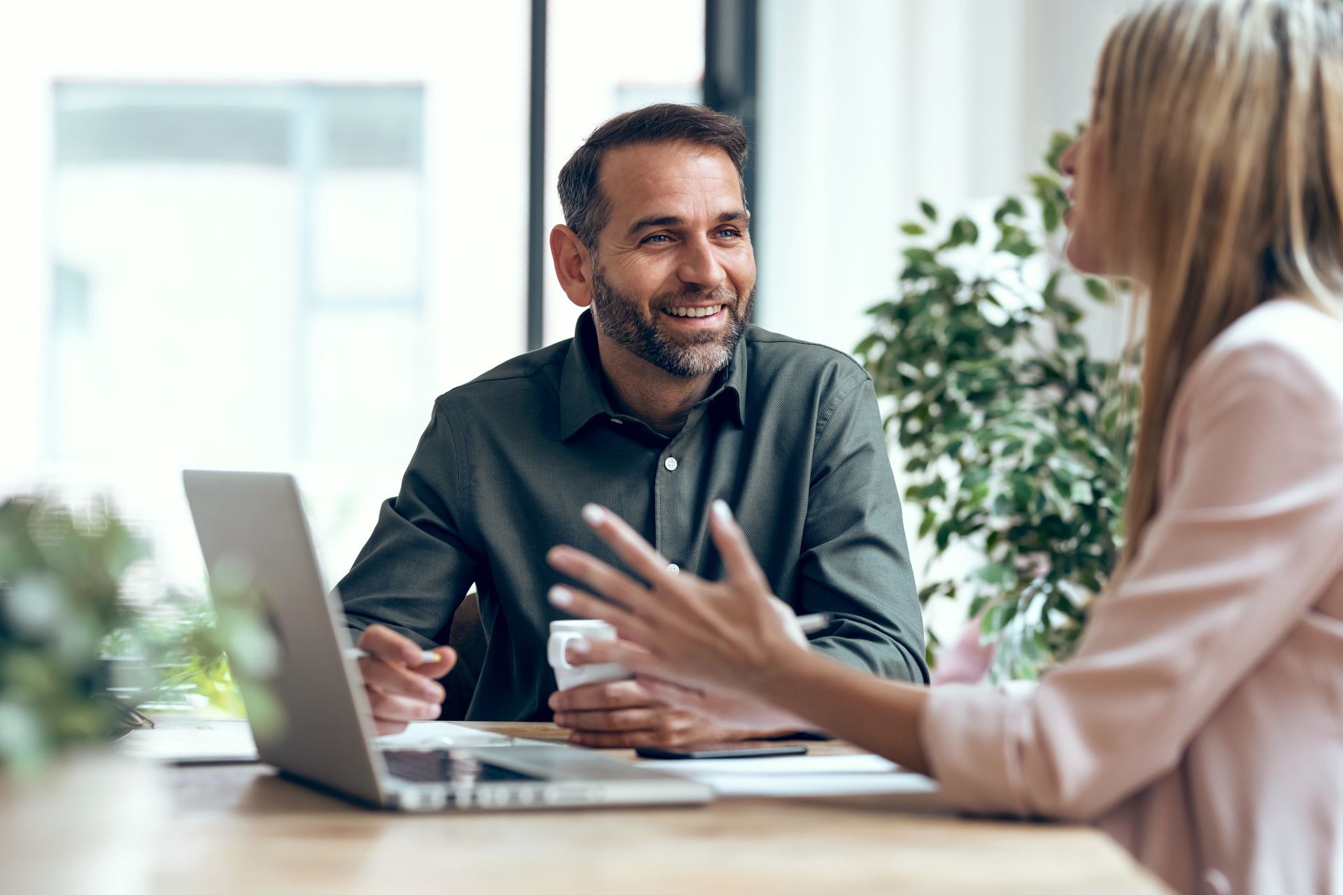 Man and woman at a table with laptop, discussing. Man smiling, holding mug. Green plants in background.
