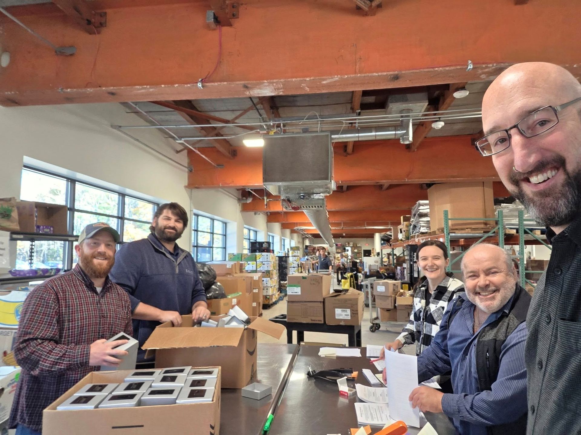 Five people packing boxes in a warehouse with an overhead crane. They are smiling and working.
