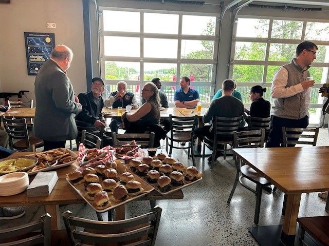 Group of people gathered indoors, eating and socializing around tables with food, near large windows.