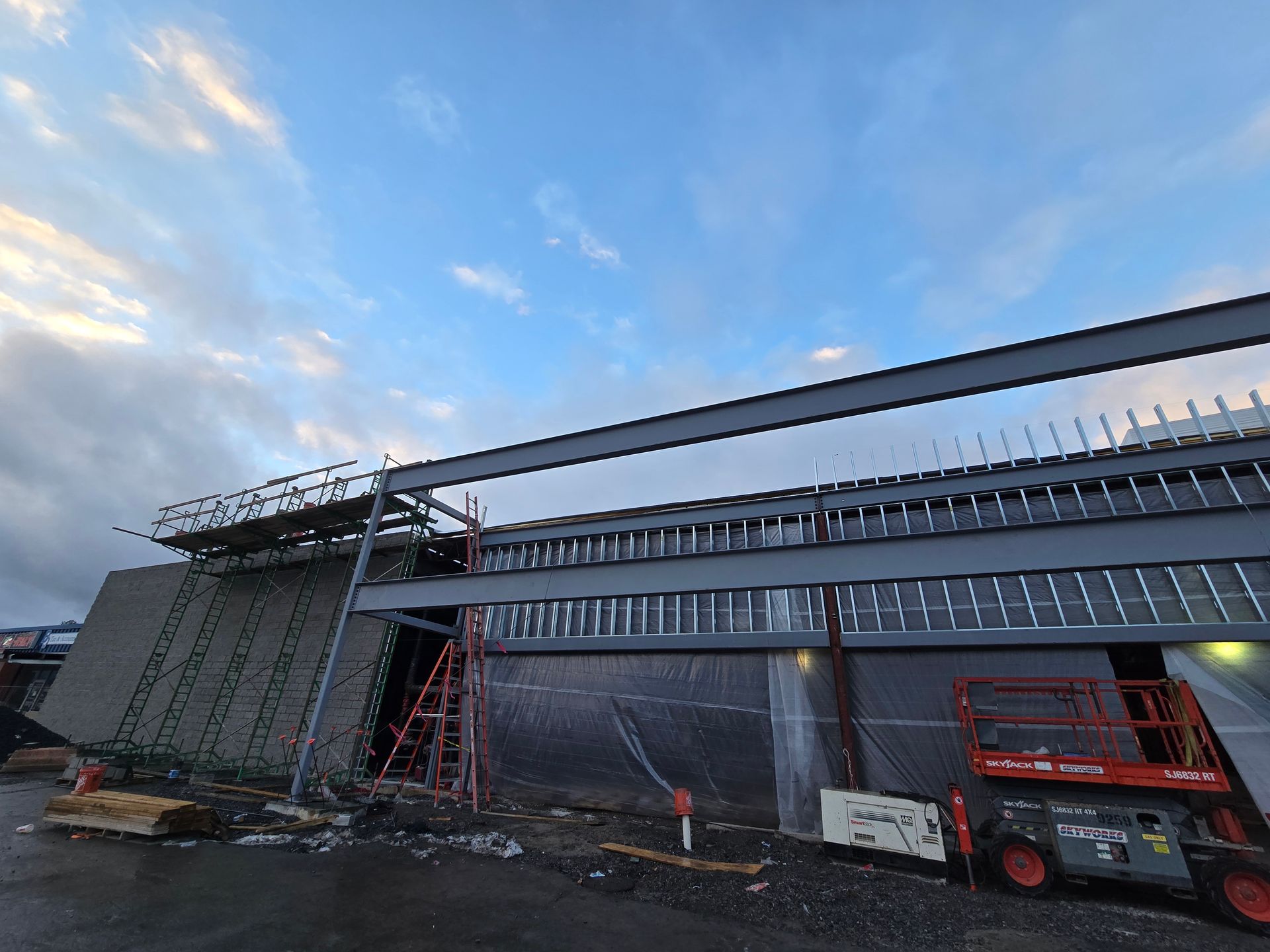 Construction site with steel beams, gray walls, and cloudy sky. A red lift and ladder are visible.