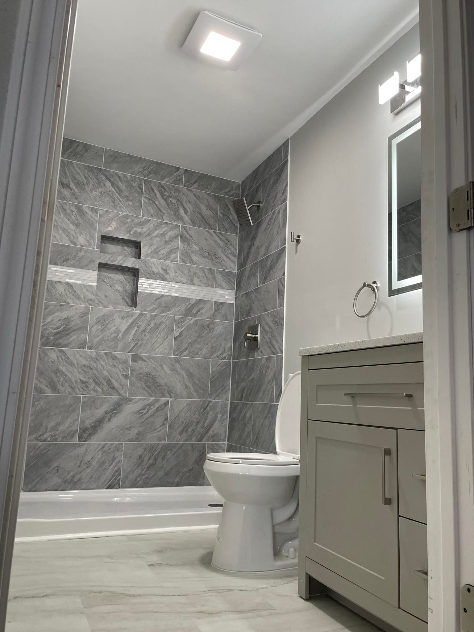 Bathroom with gray tiled shower, toilet, light gray vanity, and white walls.